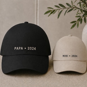Può includere: Sono mostrati due cappellini da baseball, uno nero e uno beige. Il cappellino nero ha la scritta "PAPA • 2024" stampata sul davanti. Il cappellino beige ha la scritta "MINI • 2024". Entrambi i cappellini sono su una superficie neutra.