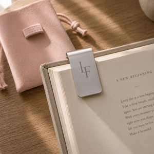 Peut inclure: Un marque-page magnétique en métal argenté gravé des lettres LF, fixé sur le haut d'une page de livre. À côté du livre se trouve une petite pochette à cordon en suédine rose clair, dotée d'une languette rectangulaire sur la face avant.