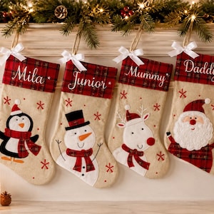 Könnte beinhalten: Vier beige Weihnachtsstrümpfe mit rot-grün karierten Bündchen, jeweils mit einer weißen Satinschleife und einem in weißer Schreibschrift gestickten Namen. Die Strümpfe zeigen applizierte Figuren: einen Pinguin, einen Schneemann, ein Rentier und den Weihnachtsmann.