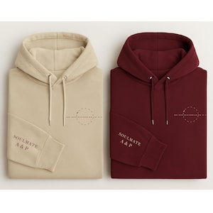 May include: Two folded hoodies, one beige and one burgundy. Each has a hood with drawstrings, a small graphic on the chest, and the text "SOULMATE A&P" on the left sleeve cuff. The beige hoodie has a dotted circle design.
