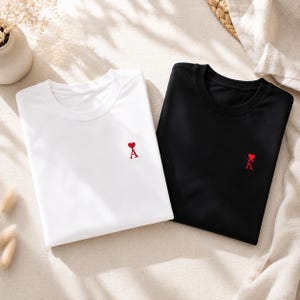 Może przedstawiać: Biały t-shirt i czarny t-shirt, oba z małym czerwonym haftem na lewej piersi. Biała koszulka posiada czerwoną literę A z sercem u góry, natomiast czarna koszulka posiada czerwoną literę K z sercem u góry.