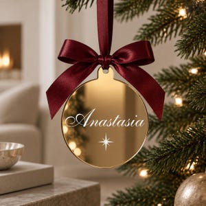 May include: A circular gold-toned mirror ornament featuring the name Anastasia in white script lettering and a small white four-pointed star icon below. The ornament is suspended by a dark burgundy satin ribbon tied in a bow at the top.