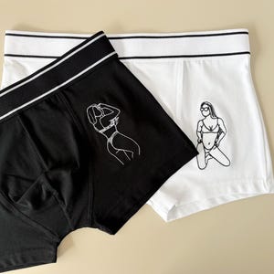 May include: A pair of underwear, one black and one white, with a graphic print of a woman in a bikini on each. The black underwear has a white waistband and the white underwear has a black waistband.
