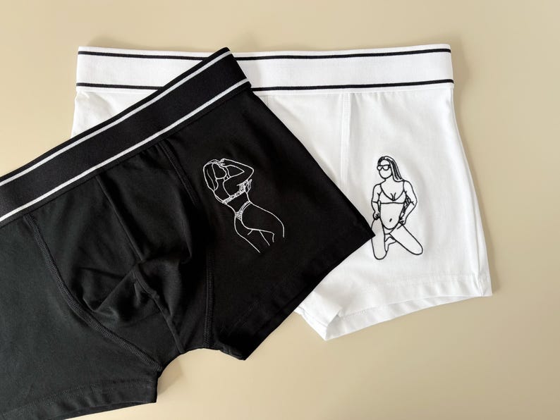 May include: A pair of underwear, one black and one white, with a graphic print of a woman in a bikini on each. The black underwear has a white waistband and the white underwear has a black waistband.