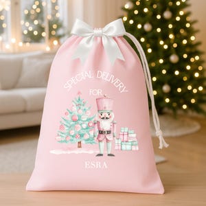 May include: A pink Christmas gift bag with a white satin bow and drawstring closure. The bag features a watercolor illustration of a Christmas tree, nutcracker, and gifts. Text on the bag reads "SPECIAL DELIVERY FOR" and "ESRA".