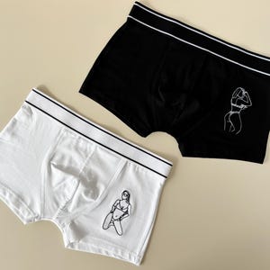 May include: Two pairs of boxer briefs with a white waistband. One pair is black with a white outline of a woman in a bikini. The other pair is white with a white outline of a woman in a bikini.
