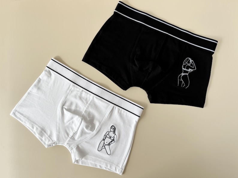 May include: Two pairs of boxer briefs with a white waistband. One pair is black with a white outline of a woman in a bikini. The other pair is white with a white outline of a woman in a bikini.