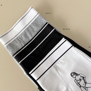 May include: Four pairs of underwear in different colors: grey, black, white, and white with a black line drawing of a woman. The underwear has a wide elastic waistband with a stripe.