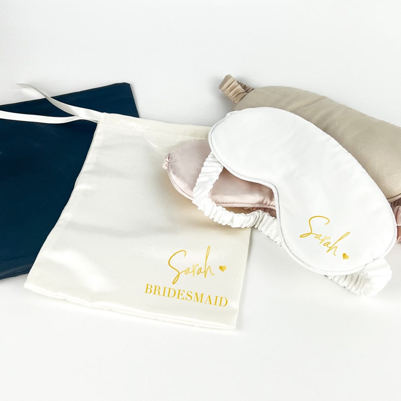 May include: A white satin sleep mask with the name "Sarah" embroidered in gold lettering and a small gold heart. The mask is in a white satin drawstring bag with the name "Sarah" and the word "BRIDESMAID" embroidered in gold lettering.