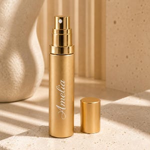 Peut inclure: Un vaporisateur de parfum cylindrique en métal doré avec un capuchon amovible assorti. Le corps du flacon présente le prénom Amelia imprimé en écriture cursive blanche. L'embout pulvérisateur est visible au sommet du récipient métallique.