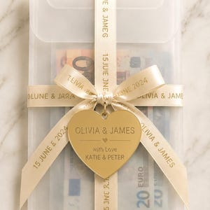 May include: A translucent rectangular envelope containing paper currency, wrapped in a cream-colored ribbon printed with "OLIVIA & JAMES" and "15 JUNE 2024". A gold-toned heart-shaped metal tag is attached, engraved with "OLIVIA & JAMES" and "with Love KATIE & PETER".