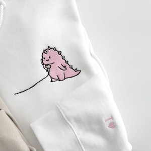 May include: White hoodie with a pink dinosaur holding a phone embroidered on the front. The hoodie also has a pink heart embroidered on the sleeve.