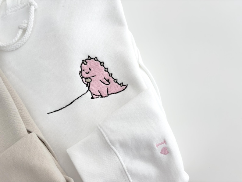 May include: White hoodie with a pink dinosaur holding a phone embroidered on the front. The hoodie also has a pink heart embroidered on the sleeve.