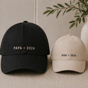 Op de afbeelding: Twee baseballpetten, één zwarte en één beige, worden getoond. Op de zwarte pet staat "PAPA • 2024". Op de beige pet staat "MINI • 2024". Beide petten staan op een neutrale achtergrond.