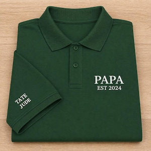 Może przedstawiać: Ciemnozielona koszulka polo z białym napisem "PAPA EST 2024" na piersi. Mankiety rękawów z białymi imionami "TATE JUDE". Koszulka ma klasyczny kołnierzyk i zapięcie na guziki.