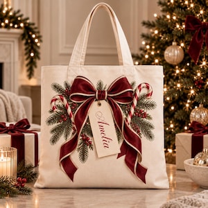 Può includere: Una borsa tote in tela color crema con un design festivo di due bastoncini di zucchero, rami di pino verdi e bacche rosse. Un grande fiocco in raso rosso scuro è centrato, con un'etichetta regalo rettangolare pendente con il nome Amelia.