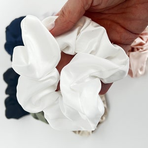 May include: A white satin scrunchie, a popular hair accessory, is held in a hand. The scrunchie is soft and has a gathered, ruffled appearance.