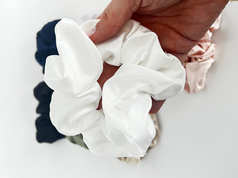 May include: A white satin scrunchie, a popular hair accessory, is held in a hand. The scrunchie is soft and has a gathered, ruffled appearance.