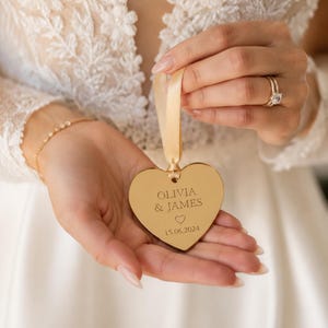 May include: A gold-toned heart-shaped ornament with a reflective surface, featuring the engraved text OLIVIA & JAMES, a small heart icon, and the date 15.06.2024. The ornament is suspended by a light beige satin ribbon loop for hanging.