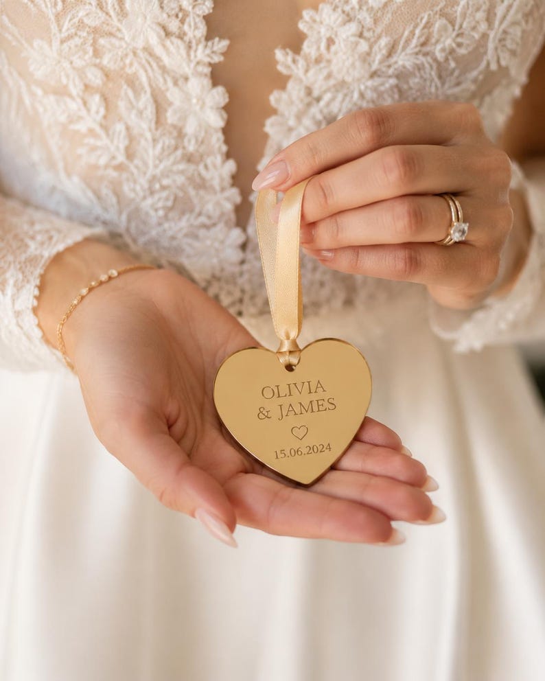 May include: A gold-toned heart-shaped ornament with a reflective surface, featuring the engraved text OLIVIA & JAMES, a small heart icon, and the date 15.06.2024. The ornament is suspended by a light beige satin ribbon loop for hanging.