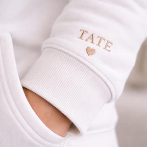 Pode incluir: Moletom branco com bolso e punhos canelados. O punho da manga apresenta o nome "TATE" bordado em fio dourado, juntamente com um pequeno s&iacute;mbolo de cora&ccedil;&atilde;o. O moletom &eacute; feito de um material macio e confort&aacute;vel.