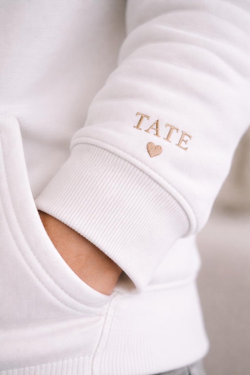 Pode incluir: Moletom branco com bolso e punhos canelados. O punho da manga apresenta o nome "TATE" bordado em fio dourado, juntamente com um pequeno s&iacute;mbolo de cora&ccedil;&atilde;o. O moletom &eacute; feito de um material macio e confort&aacute;vel.