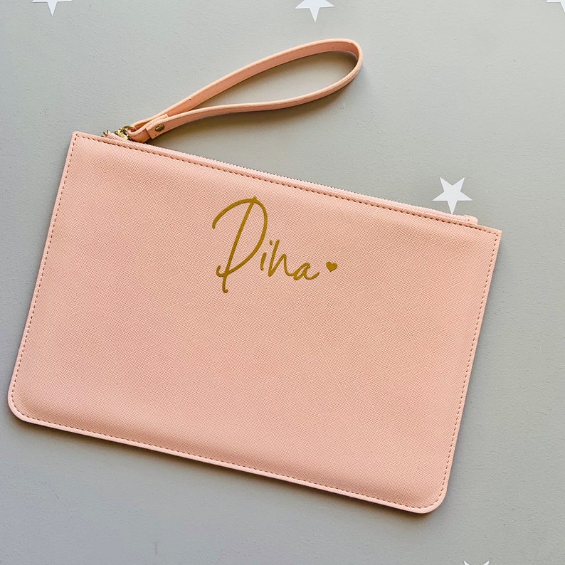 Peut inclure: Une pochette zipp&eacute;e rose p&acirc;le avec une bandouli&egrave;re dor&eacute;e et le nom "Diha" &eacute;crit en lettres dor&eacute;es avec un petit c&oelig;ur dor&eacute;.