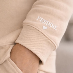 Pode incluir: Manga de camisola bege com o nome "FREDDIE" bordado em branco, acompanhado por um pequeno cora&ccedil;&atilde;o branco. A manga faz parte de um conjunto a condizer, com um bolso vis&iacute;vel. O design &eacute; simples e confort&aacute;vel.