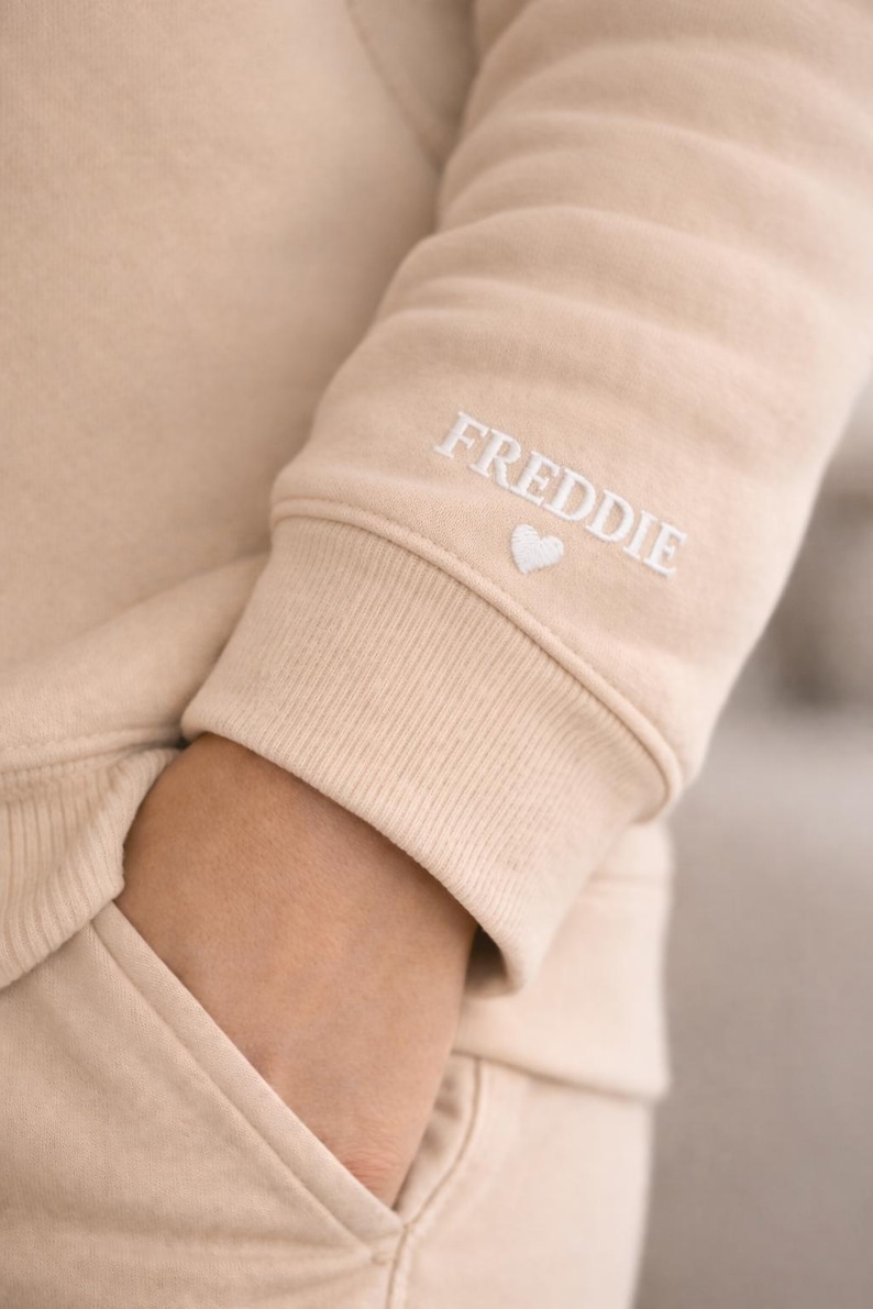 Pode incluir: Manga de camisola bege com o nome "FREDDIE" bordado em branco, acompanhado por um pequeno cora&ccedil;&atilde;o branco. A manga faz parte de um conjunto a condizer, com um bolso vis&iacute;vel. O design &eacute; simples e confort&aacute;vel.