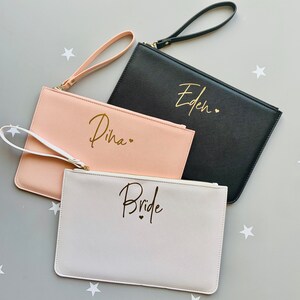 Peut inclure: Trois pochettes avec fermetures &eacute;clair et poign&eacute;es de poignet. Les pochettes sont en noir, blanc et rose. Les pochettes sont personnalis&eacute;es avec les noms "Eden", "Dina" et "Bride" en lettres dor&eacute;es.