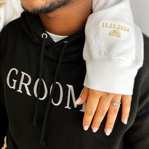 May include: A black hoodie with the word "GROOM" embroidered in white on the front. The hoodie is worn by a person with a white hoodie on top, with the date "11.11.2024" and two gold rings embroidered on the sleeve.