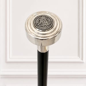 May include: A walking stick featuring a polished silver-tone circular handle with a raised, dark grey metallic center medallion. The medallion displays an intricate, embossed monogram design topped with a small crown. The shaft is solid black.