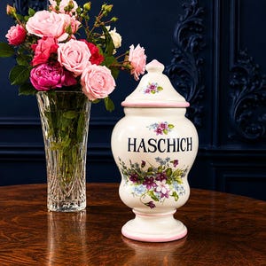 Peut inclure: Un pot en céramique blanc avec un couvercle, orné de motifs floraux et du mot "HASCHICH" en noir. Un bouquet de roses roses dans un vase en verre transparent est à côté. Le pot a un bord et une base roses, sur une table en bois.