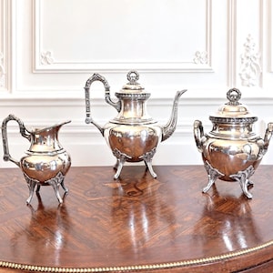 Op de afbeelding: Een driedelige theeset van zilver, bestaande uit een theepot, een melkkan en een suikerpot met deksel. Elk stuk heeft een gepolijste metalen afwerking, versierde handvatten en drie decoratieve pootjes met gedetailleerde afwerkingen.