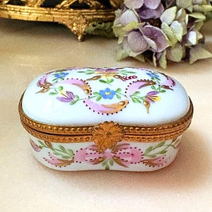 May include: An oval porcelain trinket box with a white base, featuring hand-painted pink, blue, and green floral motifs. The lid and base are joined by a gold-toned metal hinge with a flower-shaped clasp. The word Piro is written in red.