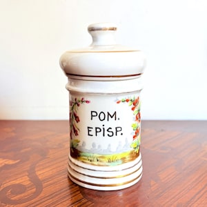 Peut inclure: Un pot d'apothicaire en céramique blanche avec couvercle assorti, orné de liserés dorés sur le bord et la base. L'avant présente le texte noir "POM. EPISP." entouré de motifs floraux peints à la main en rouge, vert et jaune, avec un paysage.