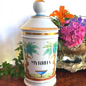 Peut inclure: Un pot à pharmacie en céramique blanche avec un couvercle, des bordures dorées et un bouton doré. Le pot est décoré de palmiers et du mot "MYRRHA". Le pot est présenté sur une surface en bois avec des fleurs en arrière-plan.