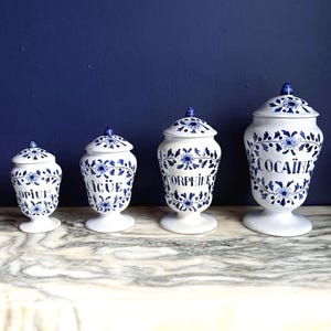 Op de afbeelding: Vier witte keramische apothekerspotten met blauwe bloemmotieven en deksels. De potten zijn gelabeld met OPIUM, IGUË, MORPHINE en COCAÏNE in zwarte letters. Elke pot heeft een voetstuk en een bijpassend afneembaar deksel met een blauwe knop.