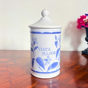 Peut inclure: Un pot en céramique blanche avec couvercle, orné de motifs floraux peints à la main en bleu et portant l'inscription "VINCA MAJOR" en lettres bleues. Le récipient cylindrique présente des bandes bleues horizontales en haut et en bas.