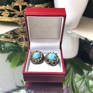 May include: A pair of turquoise and gold earrings in a red box. The box is labeled "Bijouterie Comptoir de Paris C.G. Dora Massy".