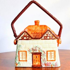 Può includere: Una teiera in ceramica a forma di cottage con facciata in mattoni color crema, tetto arancione e manico marrone. L'esterno presenta dettagli dipinti di una porta centrale, due finestre e rampicanti fioriti sulla parete anteriore.
