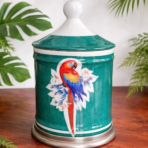 Könnte beinhalten: Eine türkisfarbene Keramikdose mit weißem Deckel und silbernem Sockel. Die Dose zeigt eine farbenfrohe Papageienillustration mit roten, gelben und blauen Federn. Die Dose ist mit weißen und blauen floralen Akzenten verziert und ist ein dekorativer Aufbewahrungsbehälter.