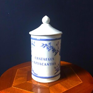 May include: A white ceramic apothecary jar with a lid, adorned with blue floral patterns and the text "CRATAEGUS OXYACANTHA." The jar sits on a wooden table with a dark blue background. The jar is approximately 6 inches tall.