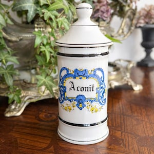 Può includere: Un barattolo da farmacia in ceramica bianca con coperchio, rifiniture nere e un'etichetta con la parola "Aconit" in un design decorativo blu e giallo. Il barattolo è cilindrico e poggia su una superficie di legno.