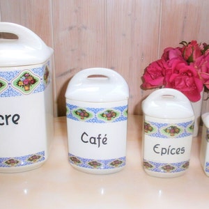 Peut inclure: Ensemble de quatre pots en céramique blanche avec des motifs floraux bleus et verts. Les pots sont étiquetés "Sucre", "Café", "Épices" et "Thé".