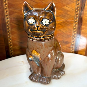 Peut inclure: Figurine de chat en céramique marron avec un papillon. Le chat a des yeux blancs et un collier. La figurine est vernissée et a une finition brillante. Le chat est assis droit, les pattes devant.