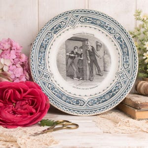 Peut inclure: Assiette ancienne en porcelaine avec une illustration centrale en noir et blanc d'une scène avec des personnages et du texte. L'assiette a une bordure florale bleue et blanche, entourée de fleurs et de décor vintage.