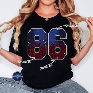 May include: Black t-shirt featuring the number "86" in a sparkling design. The number is composed of blue and red pixelated patterns, outlined with a border of small, shiny embellishments. The shirt has a classic crew neck.