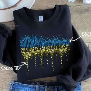 May include: Black sweatshirt with the word "Wolverines" in blue and yellow rhinestone-style lettering. The design features a dotted pattern, with blue dots at the top and yellow dots dripping down. The sweatshirt is folded on a wooden surface.