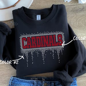 May include: Black sweatshirt with the word "CARDINALS" in red rhinestones. The word is outlined in white rhinestones, with a dripping effect. The sweatshirt is folded, with a white hat and blue jeans in the background.